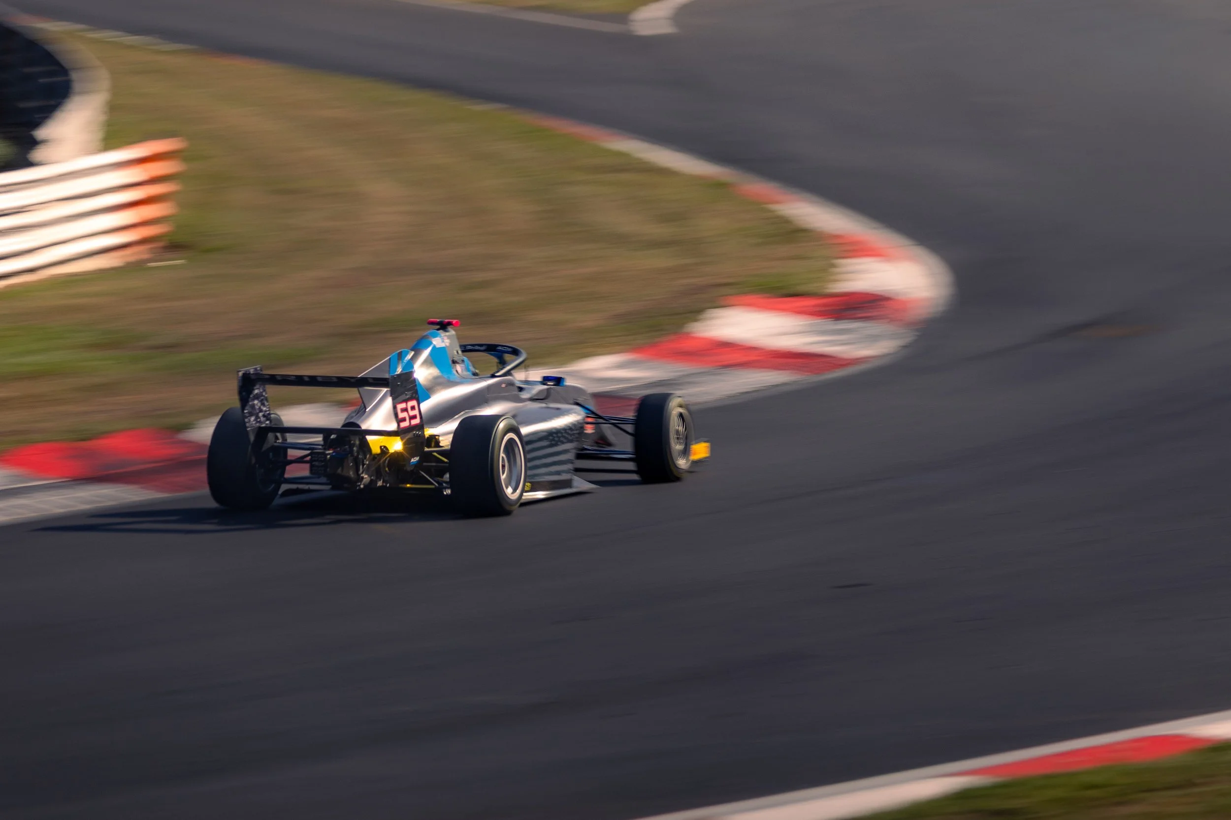 A race car with the number 59 on it is speeding around a race track curve, with blurred background and trackside grass visible.