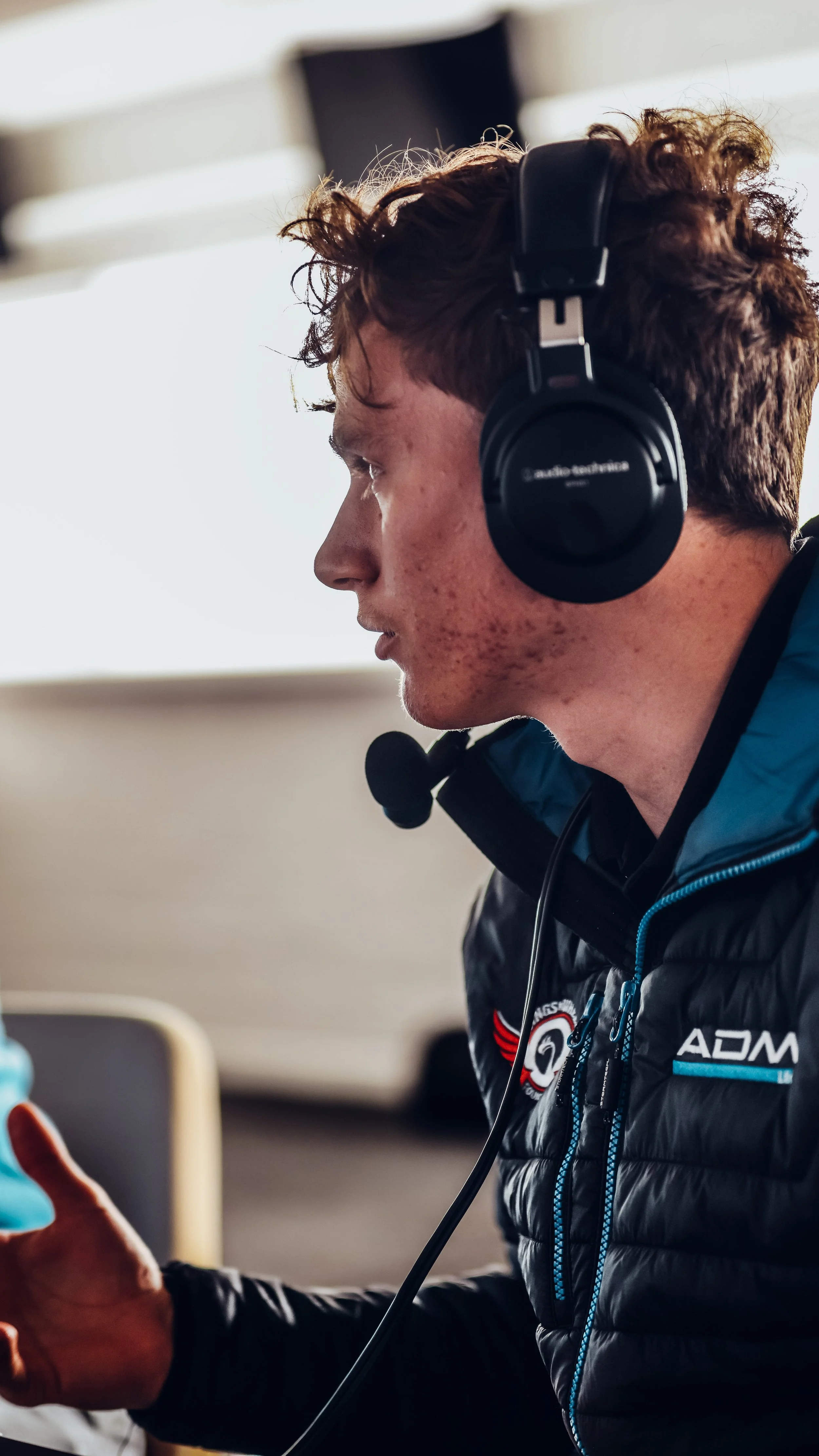A young man with curly hair wearing a black headset with a microphone, sitting at a table, looking down, possibly working or studying in an indoor setting.
