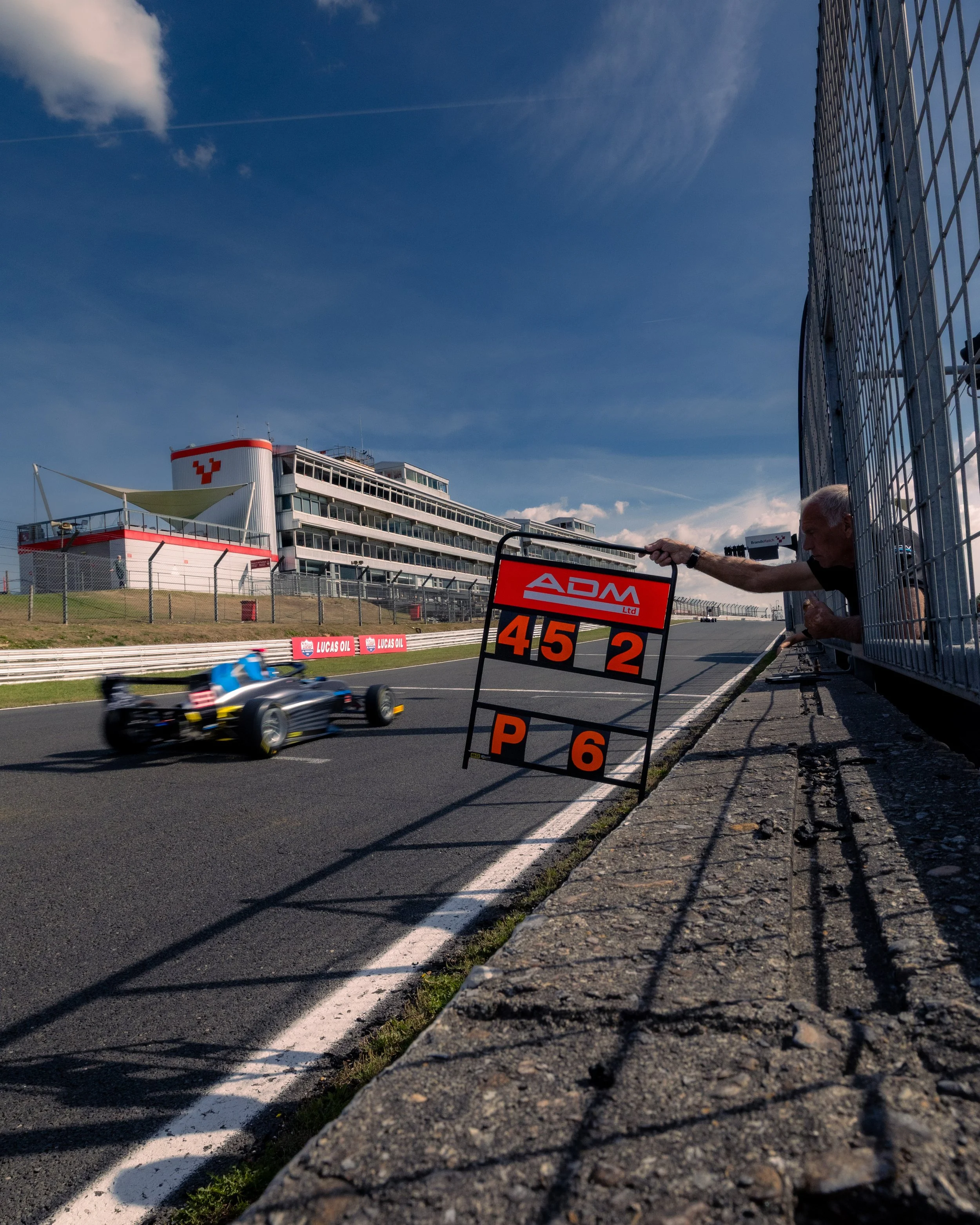 A race car goes by on a race track with a person holding a pit board displaying the lap times, under a blue sky with some clouds.