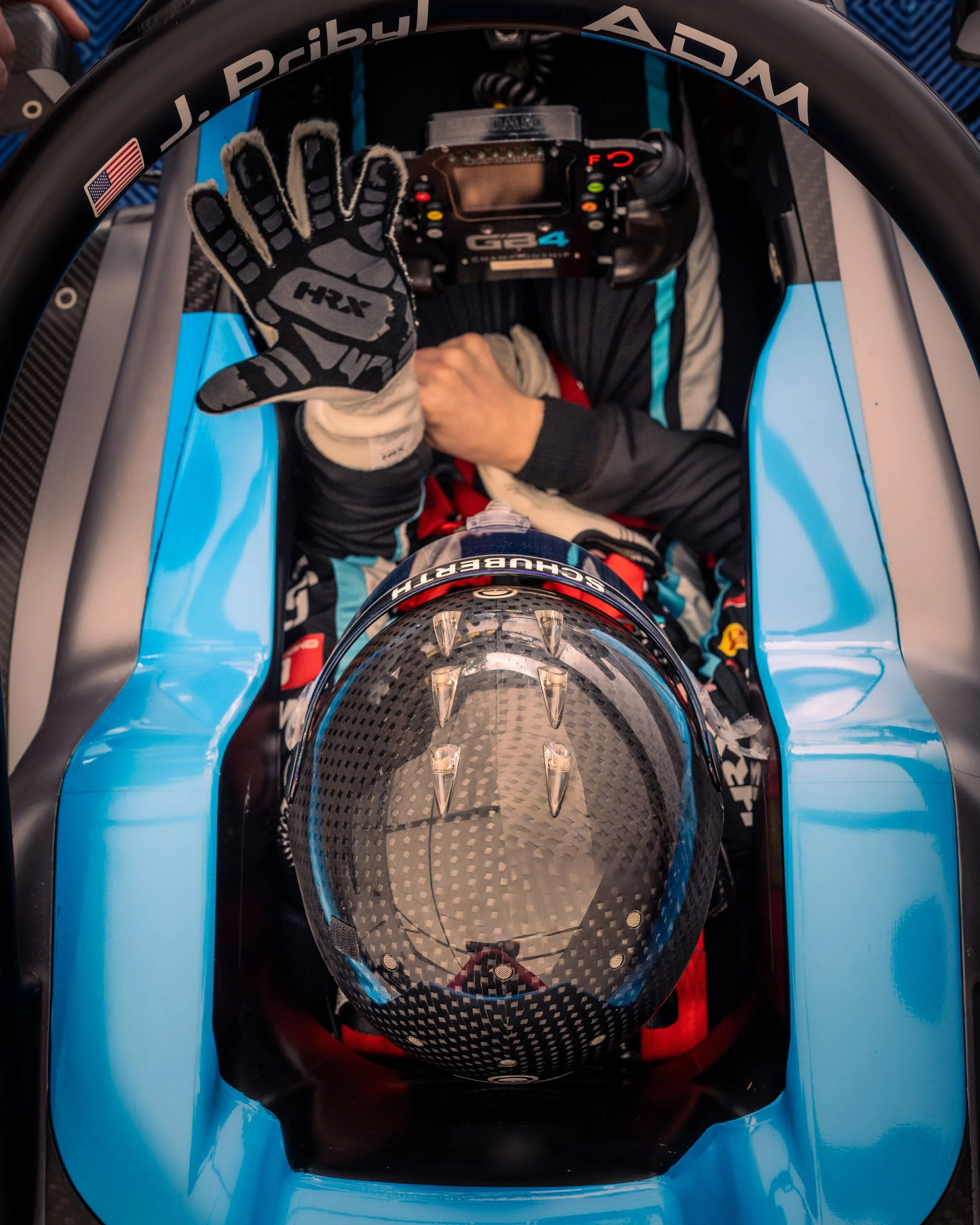 Race car driver in a cockpit, wearing a helmet with a carbon fiber pattern, and a racing suit. The driver is seated inside a blue racing car, holding a racing glove and making a peace sign with their right hand.