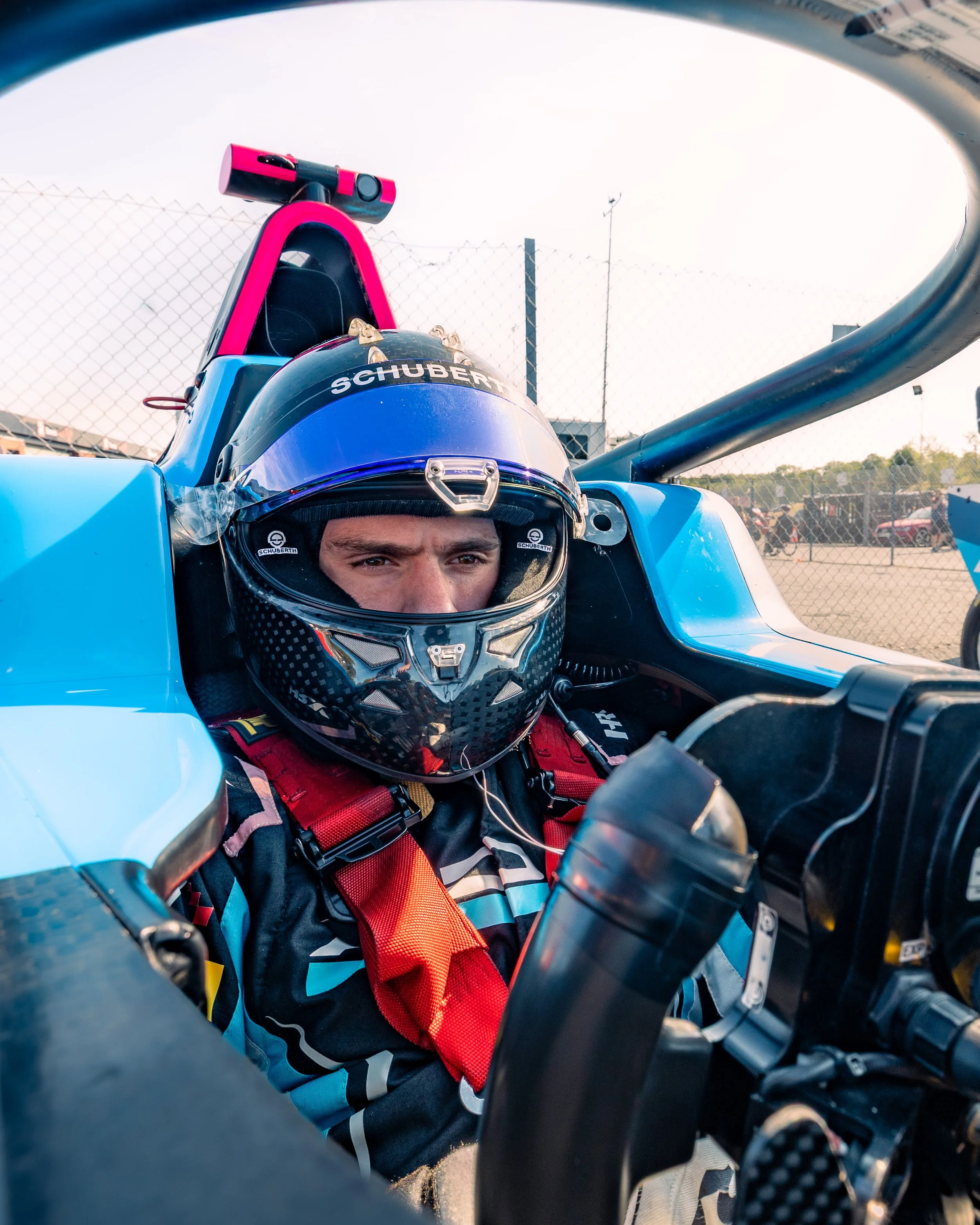 Race car driver sitting in a blue race car cockpit, wearing a black helmet with a face shield and a racing suit with red harness straps.