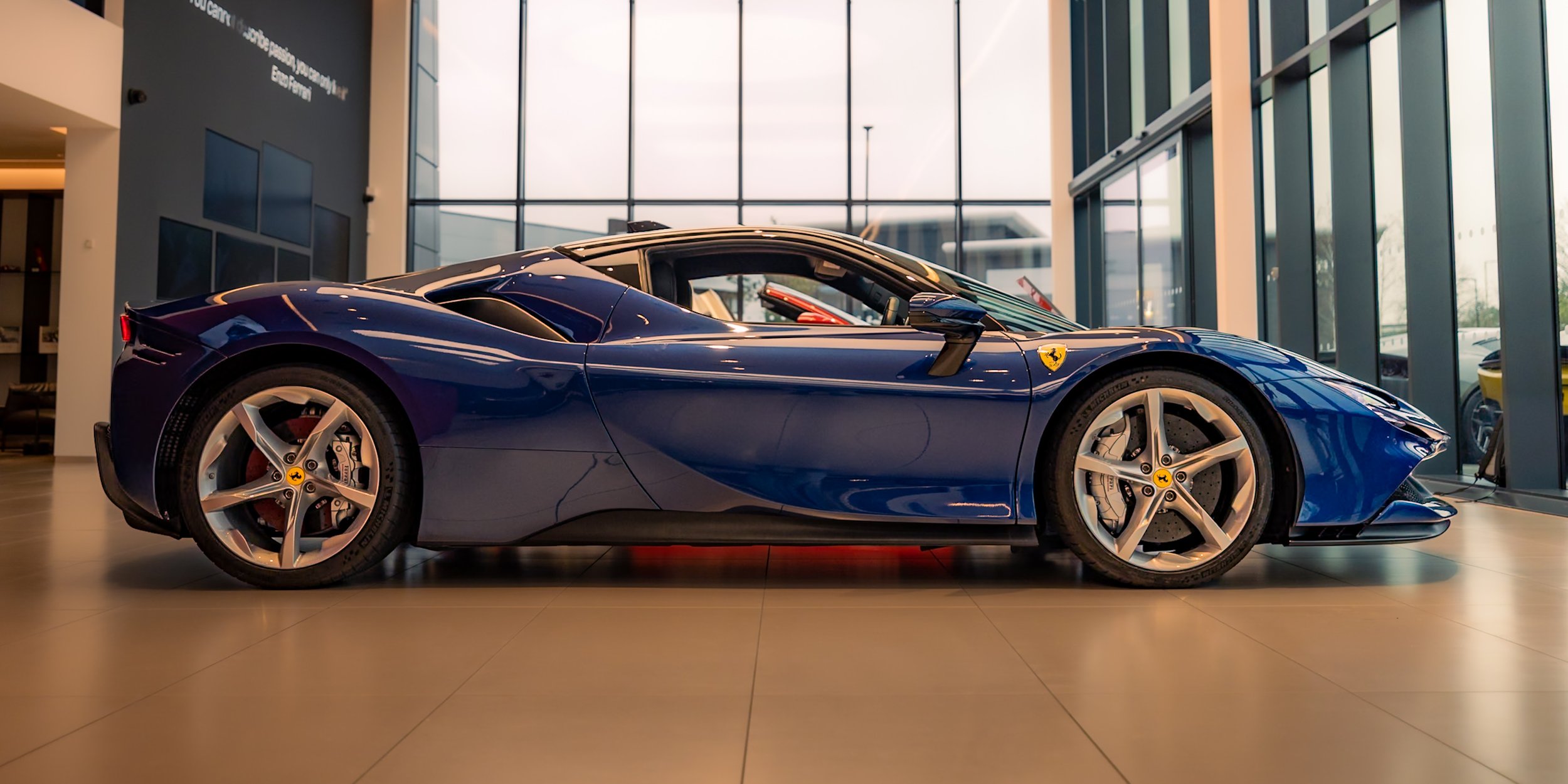 A blue Ferrari sports car displayed in a showroom with large windows.