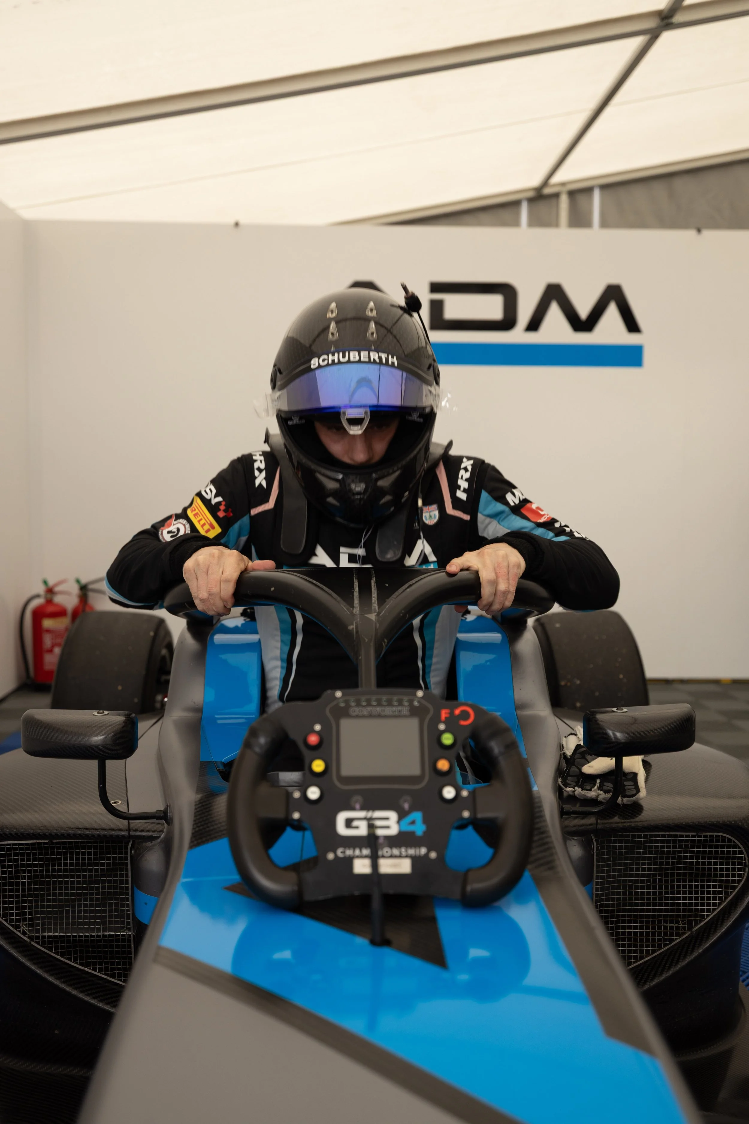Race car driver wearing a helmet and racing suit sitting in a blue Formula 4 race car, inside a racing garage with fire extinguishers in the background.