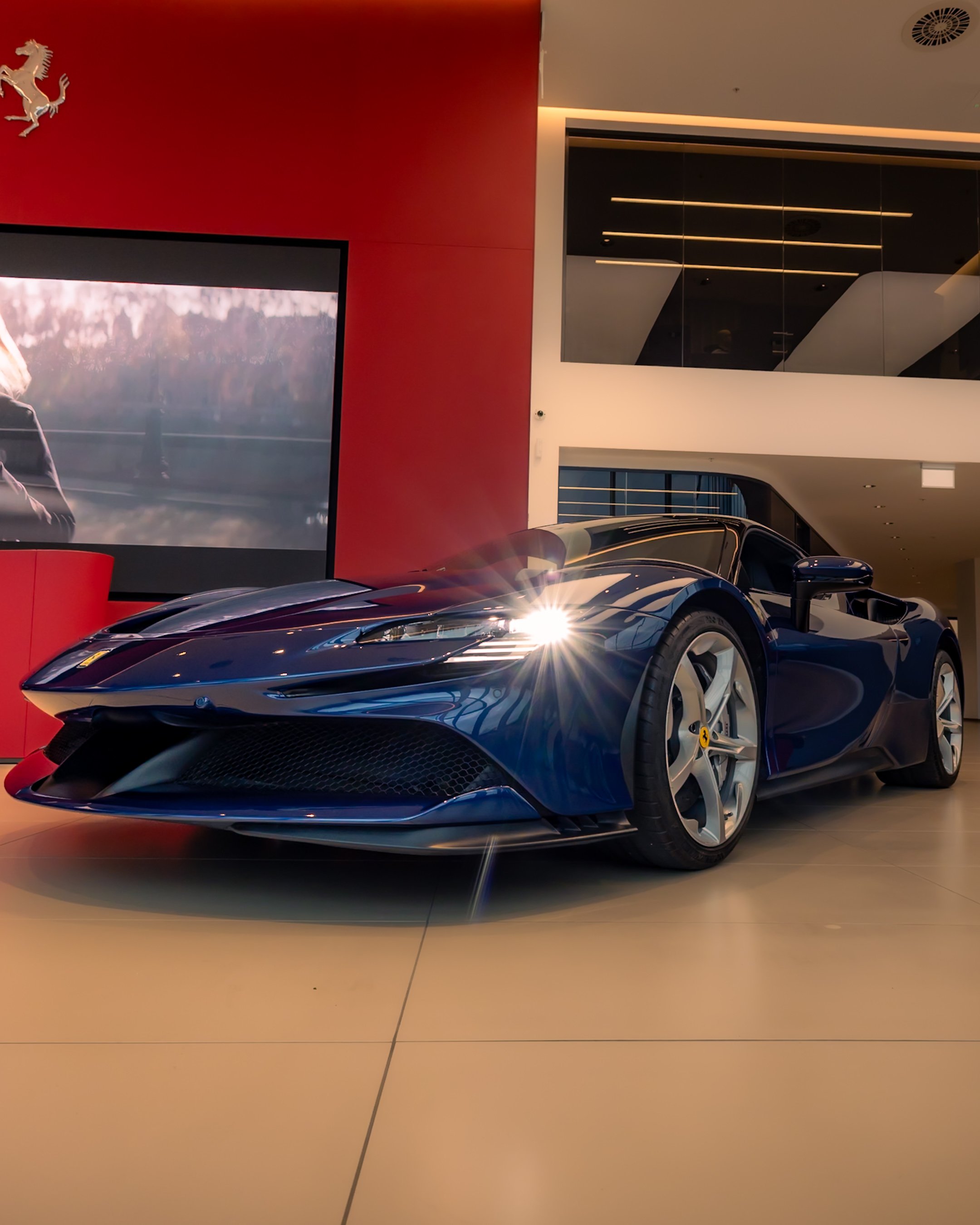 Blue sports car on display in a showroom with red and white walls, large screen in the background, and a Ferrari logo.