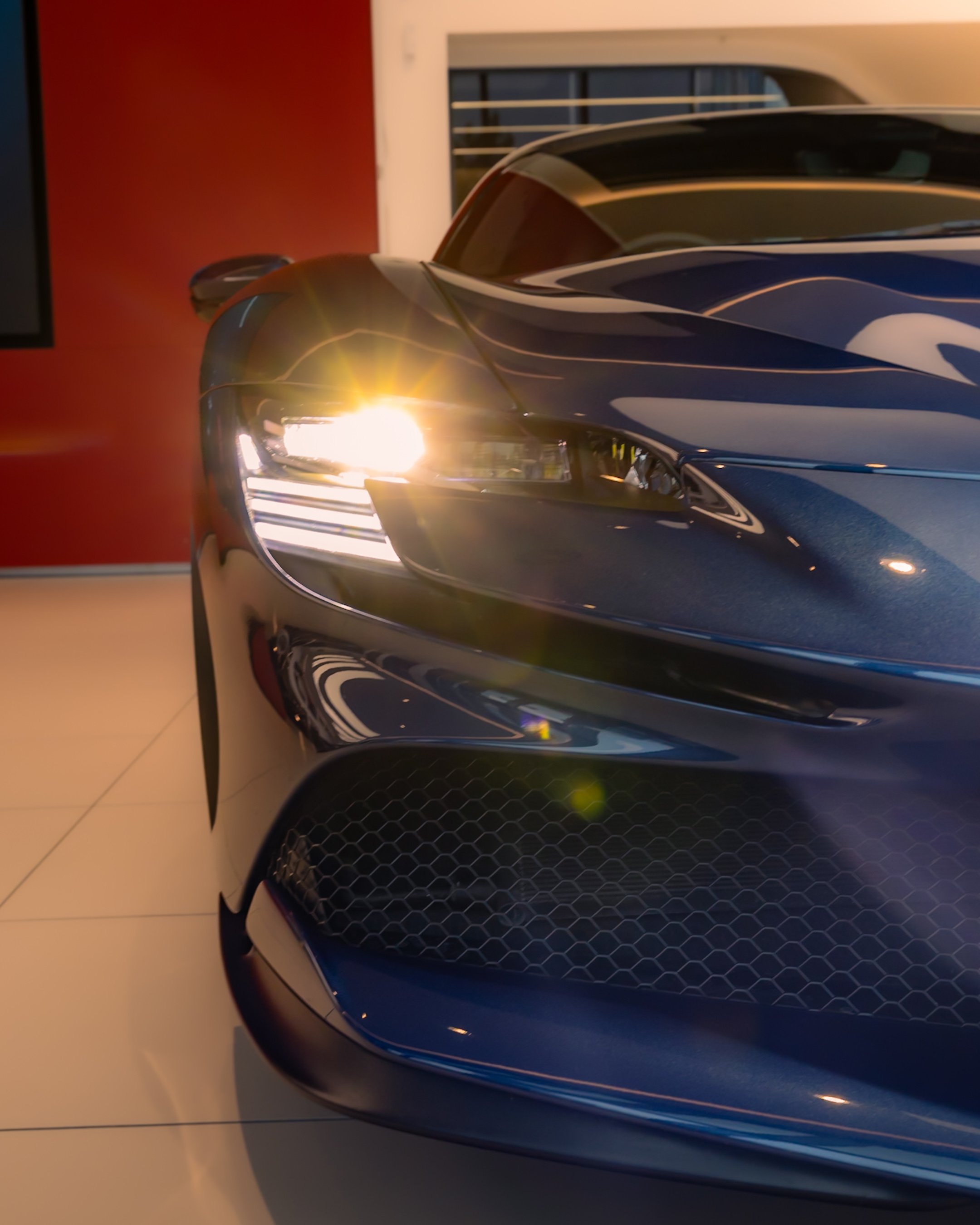 Close-up of the front headlight and grille of a sleek blue sports car with the headlight turned on.