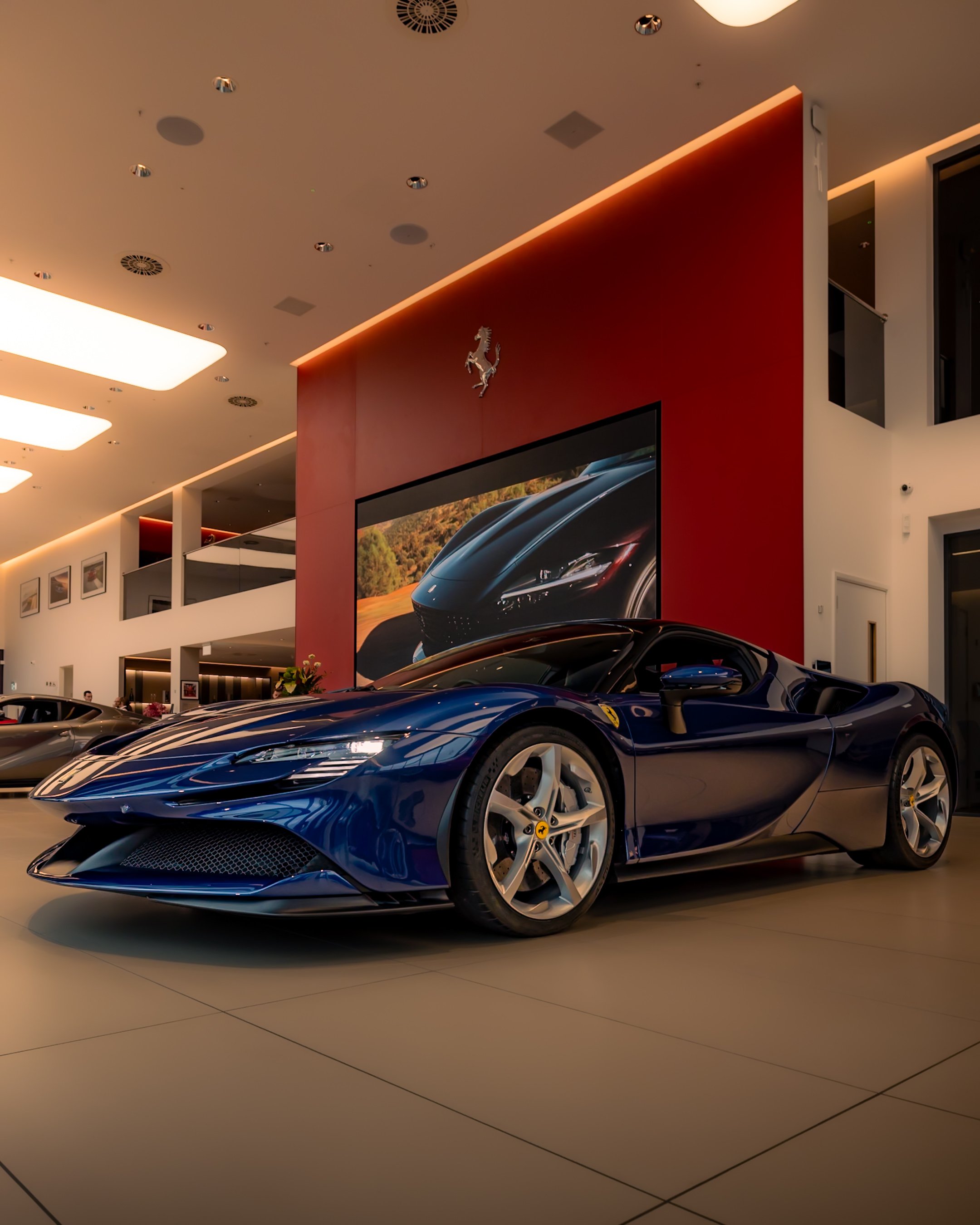 A blue sports car displayed in an upscale showroom with modern lighting and red accent walls, featuring a large screen displaying a car image.
