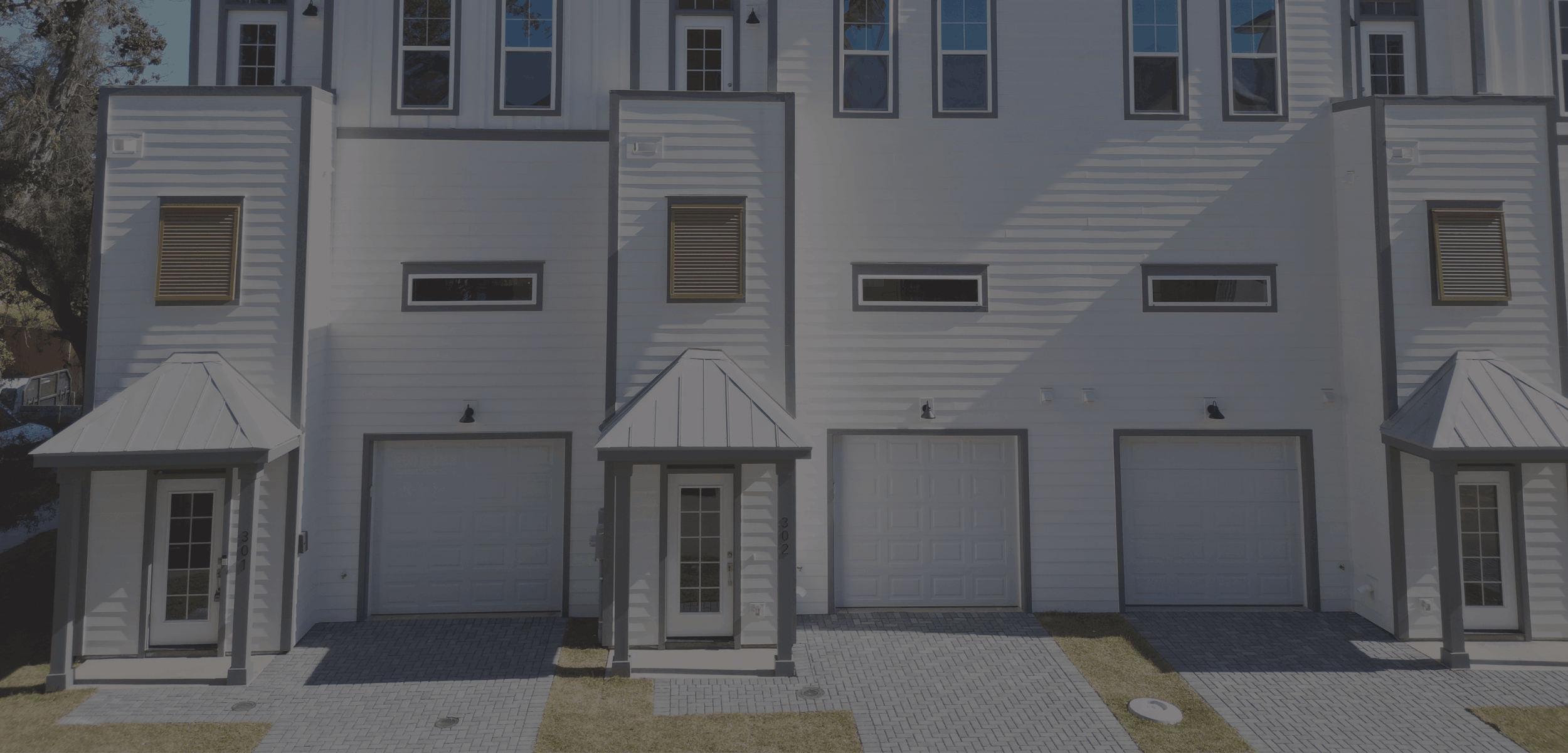 Front view of a modern white townhouse with three garage doors, small entry doors, and small windows, with paved pathway and small patches of grass in front.