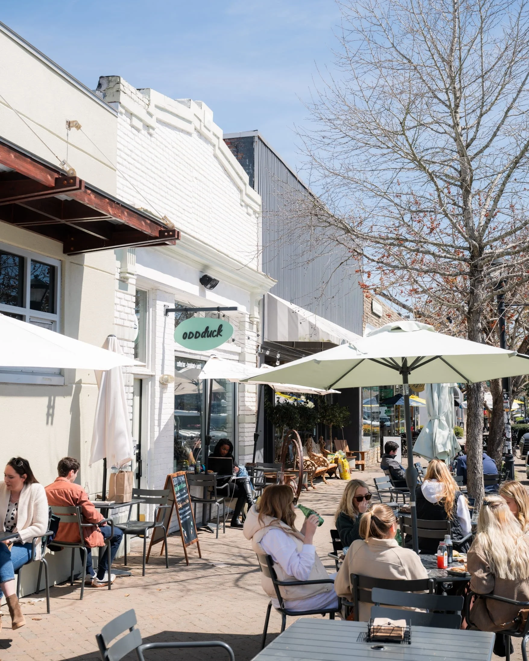 Outdoor patio scene with several people seated at tables under cream-colored umbrellas in Park Circle. There are buildings with storefronts, including a sign for 'Odd Duck', and leafless trees in the background. The sky is clear and blue.