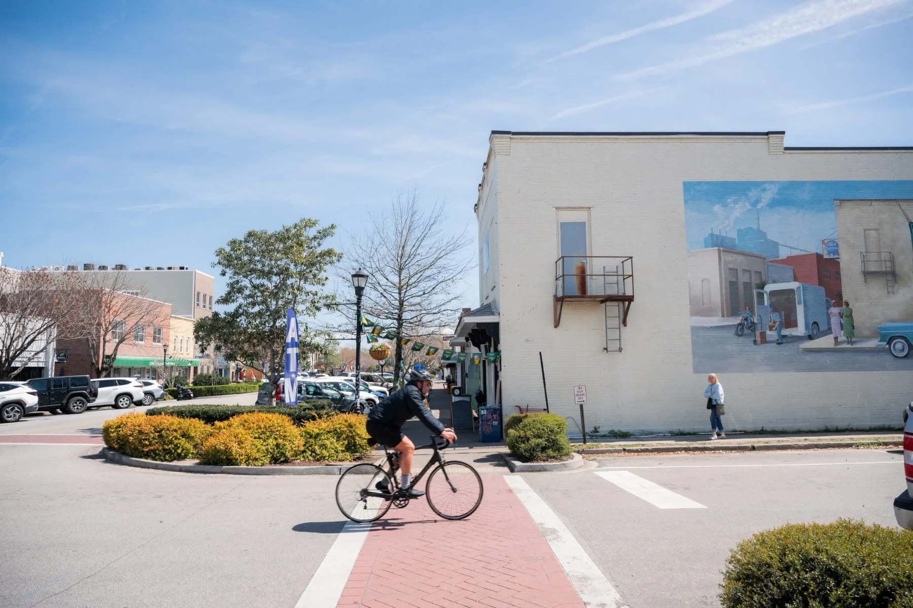 A street scene with a man riding a bicycle across a crosswalk at Park Circle in Charleston, South Carolina, a woman standing on the sidewalk, cars parked along the road, and a mural painted on a beige building wall.