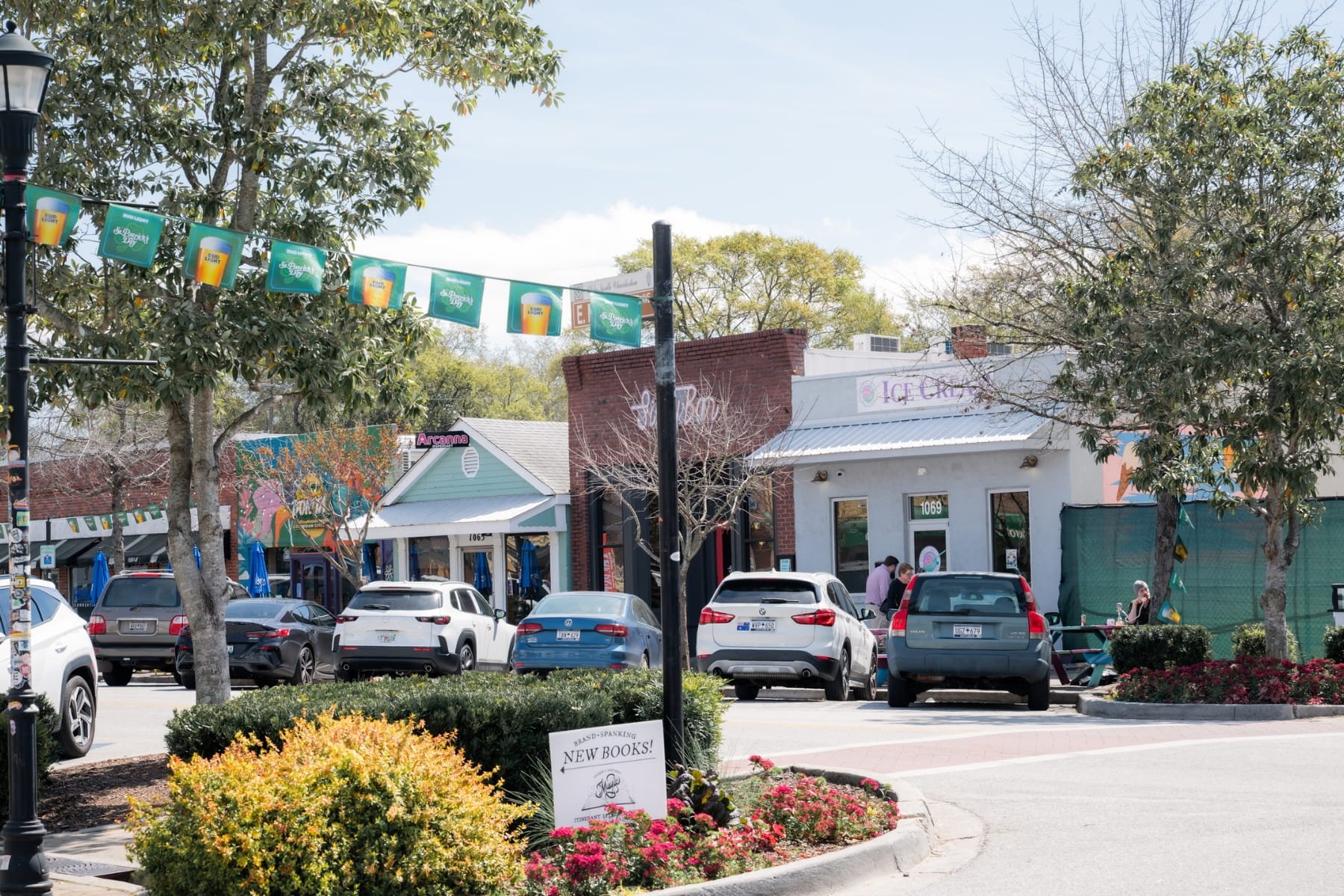 Street scene with parked cars, storefronts, trees, and holiday banners in a small shopping area