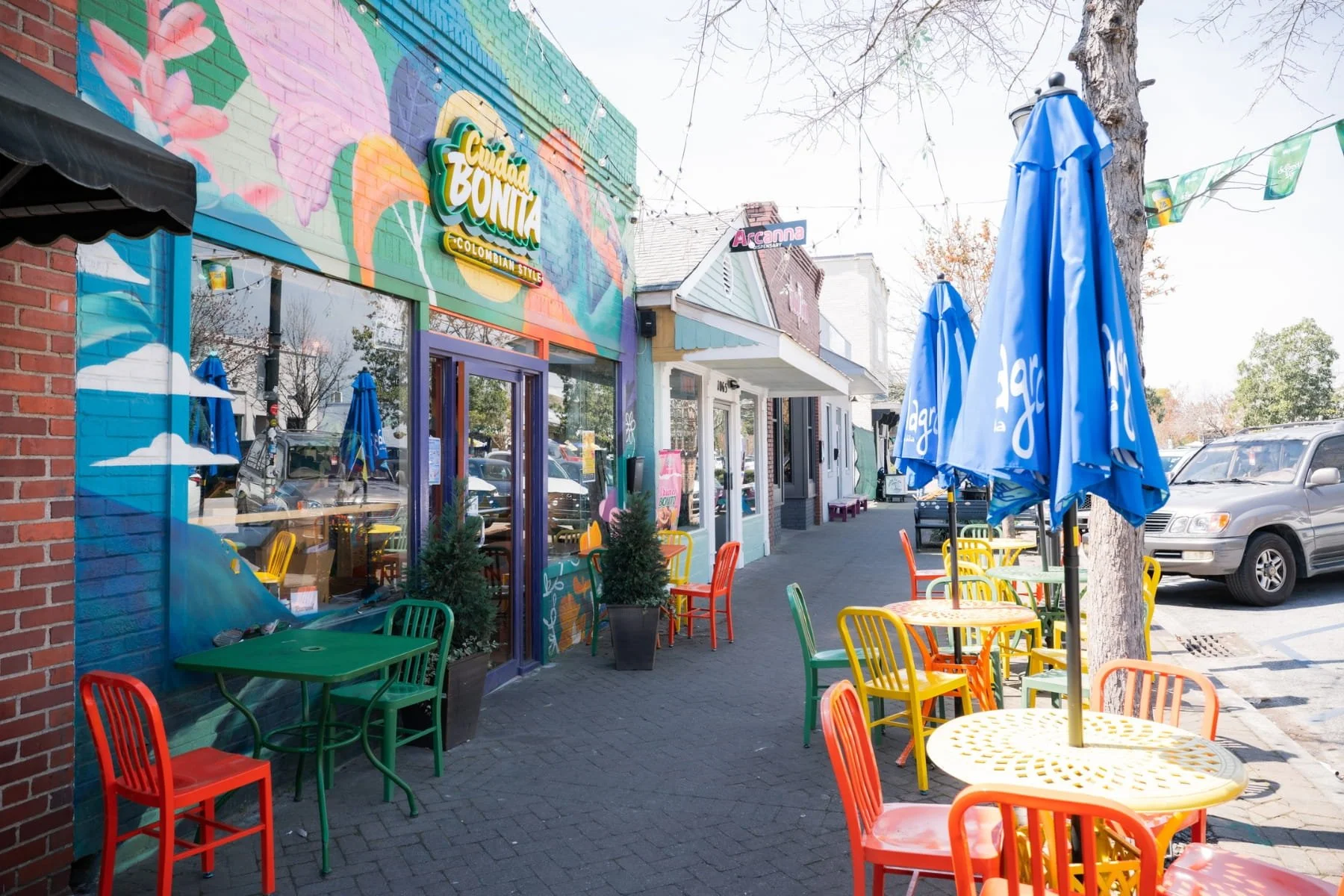 Colorful outdoor patio of a Park Circle restaurant with green, orange, yellow, and red chairs, tables with large blue umbrellas, and a mural on the building wall.