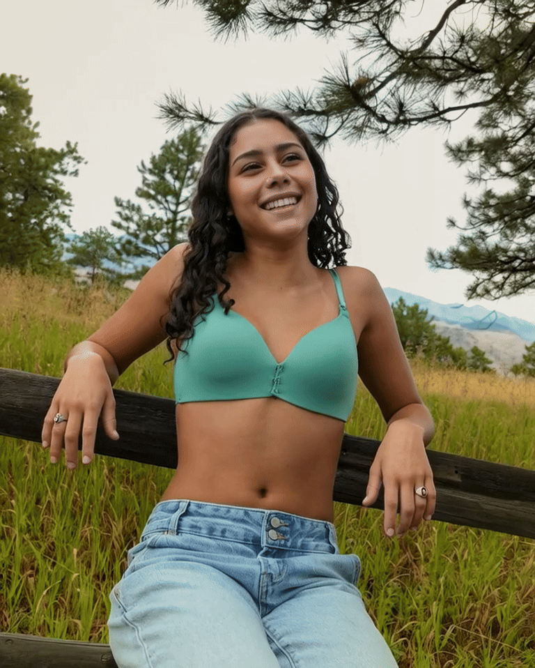 Young woman with curly dark hair smiling outdoors, sitting on a wooden fence in a grassy field with trees and mountains in the background.