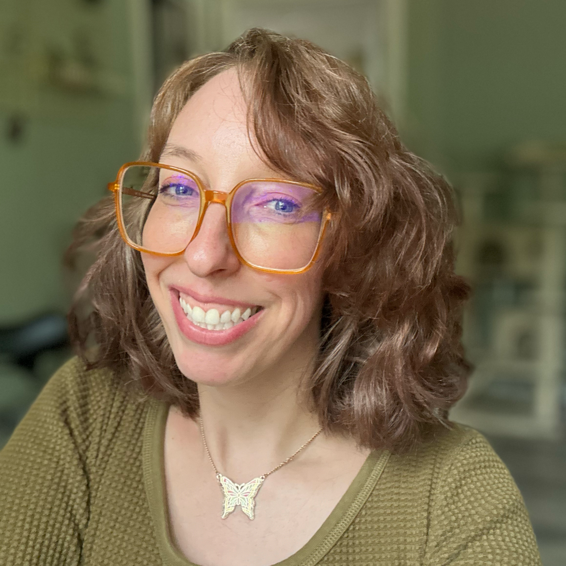 A woman with long, wavy auburn hair wearing large orange glasses and a butterfly necklace, smiling in an indoor setting.