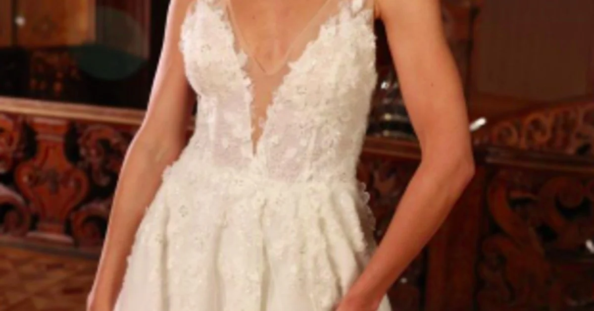 A woman wearing a white lace bridal gown standing beside carved wooden railings in an ornate interior.