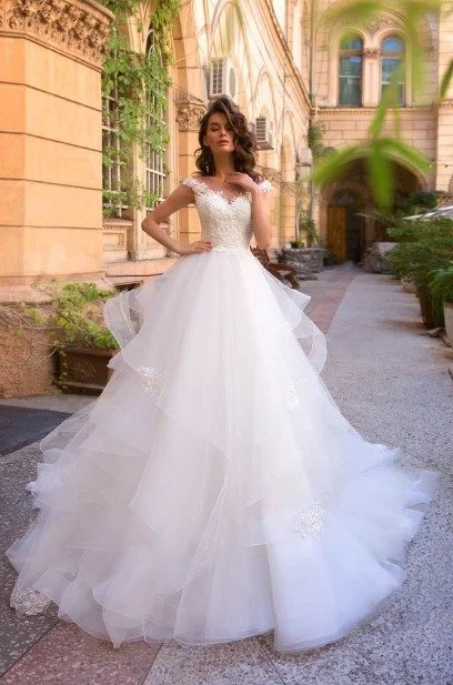 Bride in a lace bodice ball gown with a layered tulle skirt poses in an elegant courtyard beside a historic building.