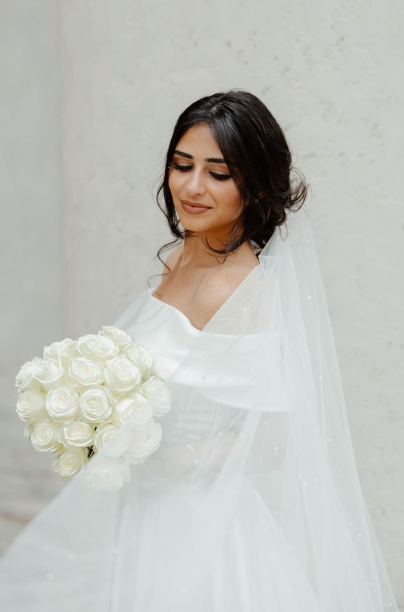 Bride in a white wedding gown and long veil holds a bouquet of white roses while looking down by a stone wall.