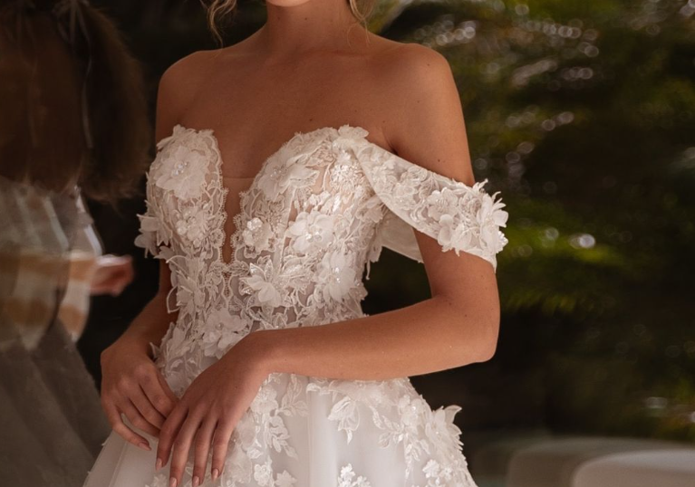 Close-up of a bride wearing an off-the-shoulder floral lace wedding gown with a fitted bodice in an outdoor setting.