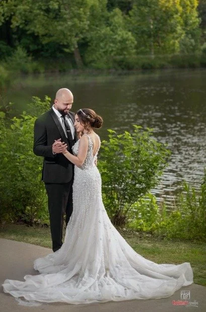 Bride in a fitted white lace gown with a long, flowing train dances with the groom beside a lake surrounded by greenery.