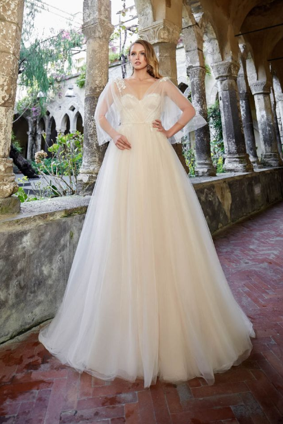 Bride in a flowing ivory wedding gown with sheer sleeves poses among stone arches in a lush historic courtyard.