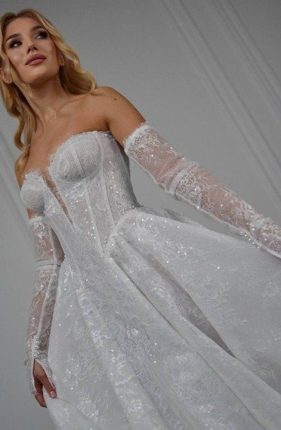 Bride wears an off-the-shoulder lace wedding gown with sheer long sleeves, a corset bodice, and a full skirt.