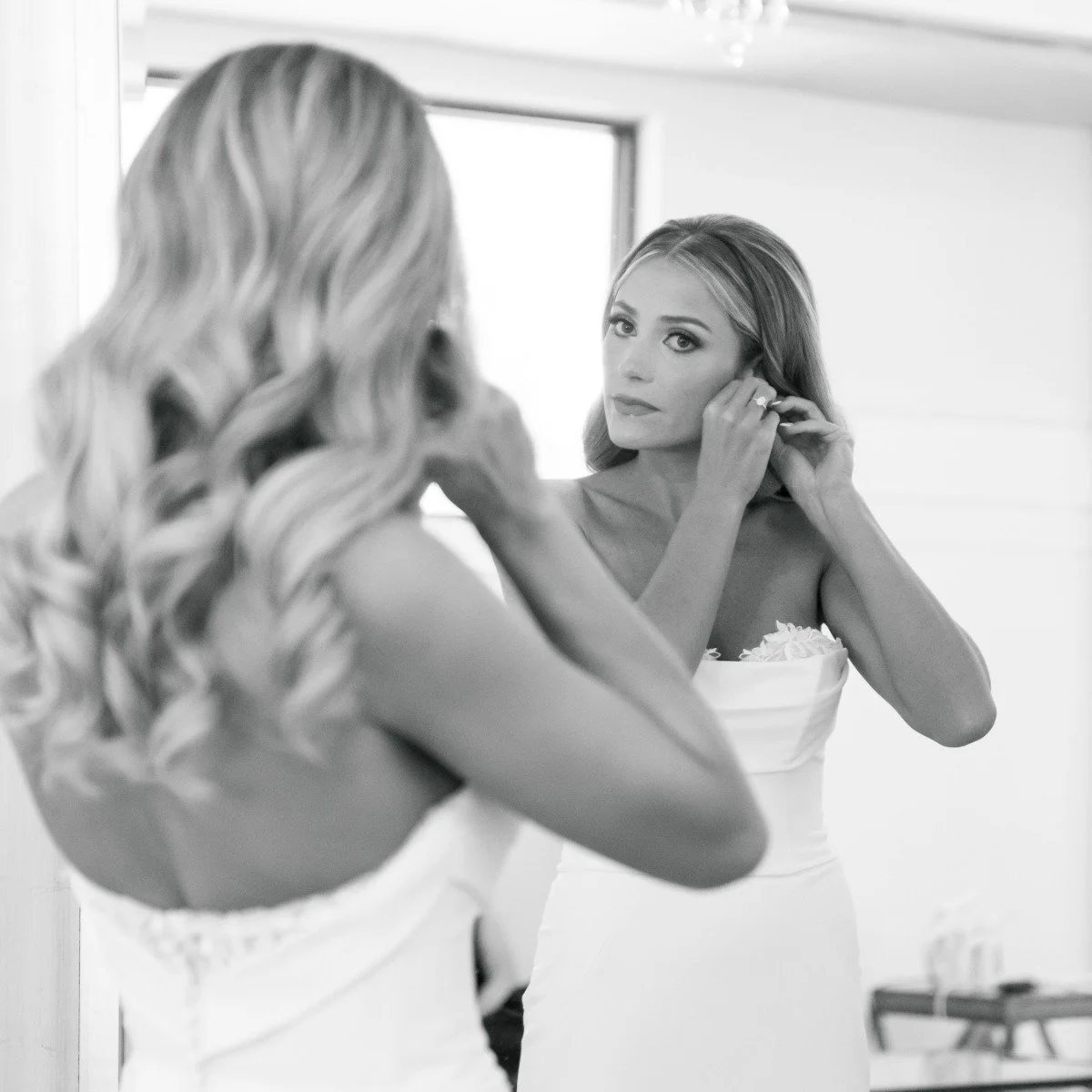 A woman in a strapless white dress adjusting an earring while looking into a mirror in a bright room.