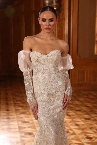 Bride in a fitted beaded wedding gown with sheer off-the-shoulder sleeves poses in an elegant wood-paneled room.