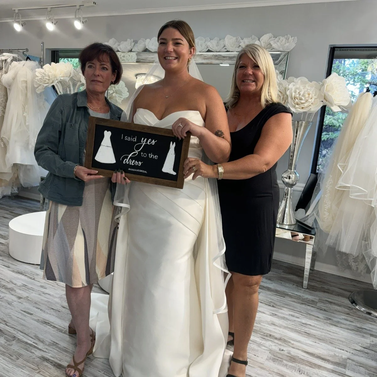 Three women standing together in a bridal shop, holding a framed sign among white dresses and large flowers.