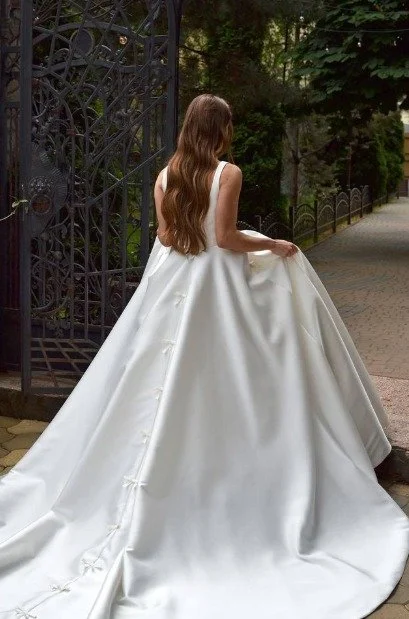 Bride with long brown hair in a white satin ball gown with a sweeping train and floral buttons beside an iron gate.