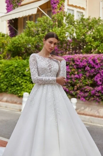 Bride in a long-sleeve lace ball gown poses outdoors in front of lush green shrubs and bright purple flowers.