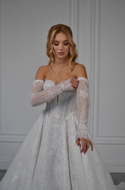 Bride in a sparkling white lace ball gown with off-the-shoulder sleeves adjusts one sleeve against a paneled wall.