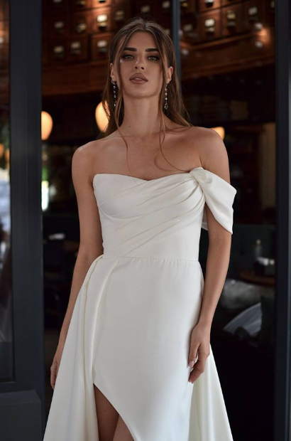 Bride wears a sleek white one-shoulder wedding gown with a draped bow, fitted waist, thigh-high slit, and flowing skirt.
