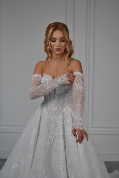 Bride in an off-the-shoulder lace wedding gown adjusts a sheer lace sleeve against a white paneled wall.