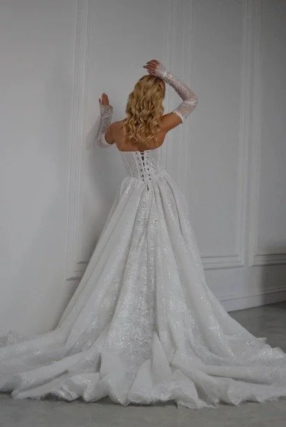 Bride with blonde curls wearing a sparkling white ball gown with a long, flowing train and sheer lace sleeves.