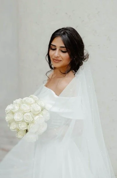 Bride in a flowing white wedding gown and veil holds a bouquet of white roses while looking down softly.