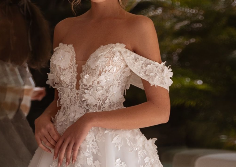 Bride wears an off-the-shoulder white lace wedding gown with floral appliqués, a plunging neckline, and fitted bodice.