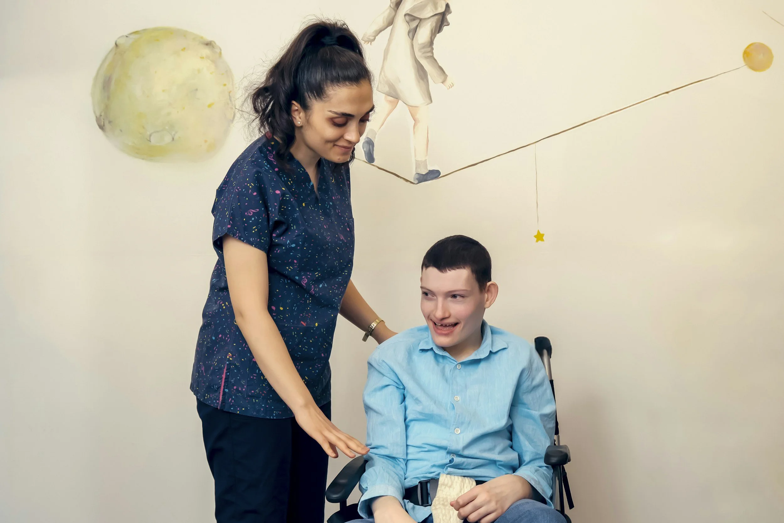 A young man with IDD sits in a wheelchair as a woman who is his caregiver smiles down at him.