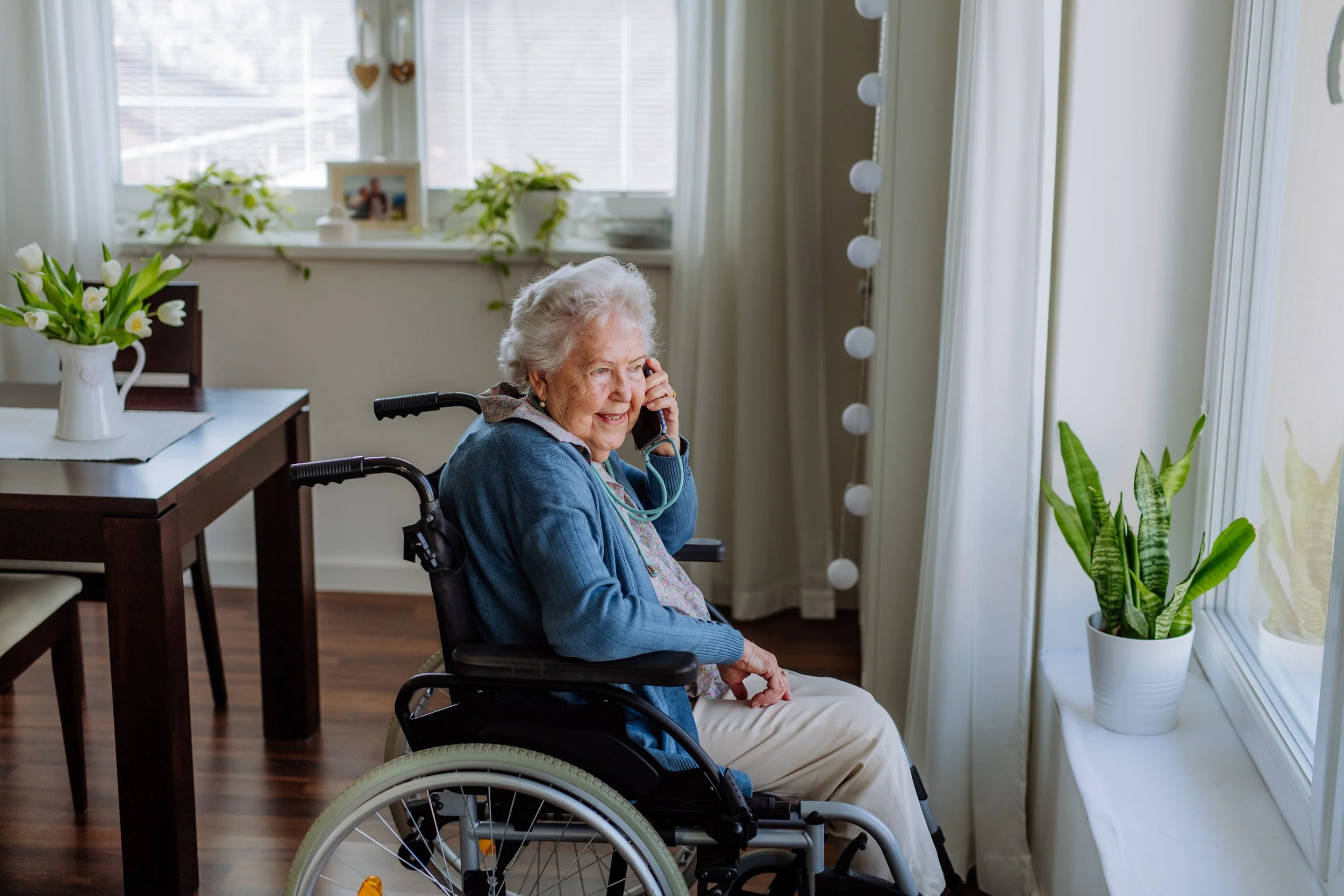 Image: An elderly woman who is a wheelchair user looks out of the window of her home while talking on the phone.