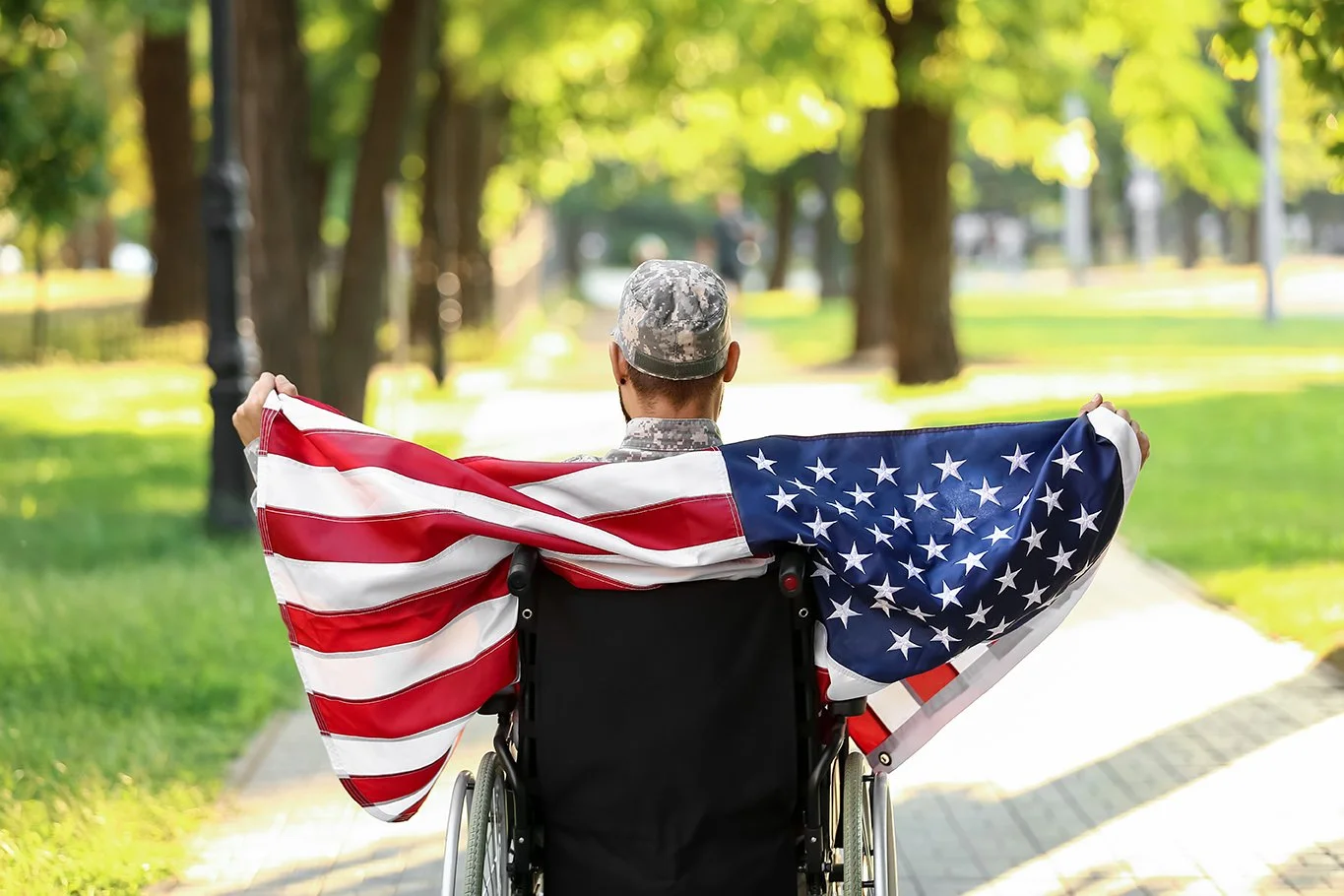 A man who is a wheelchair sits in his chair outside. He is wearing an army cap, holds an American flag behind himself.