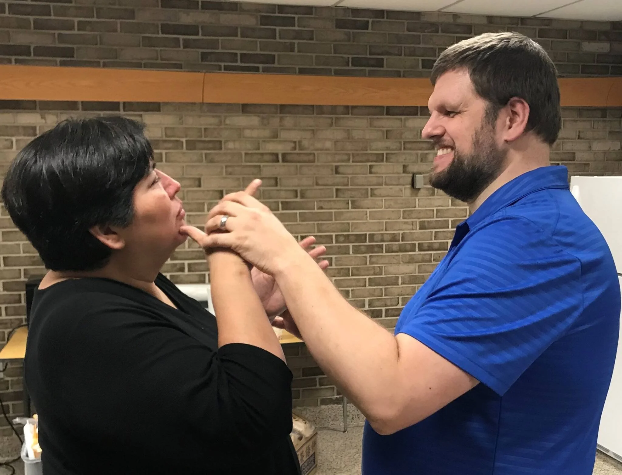 A woman and a man face each other, hands intertwined near face level as they communicate using tactile sign language. The man is smiling broadly. A brick wall is visible in the background.