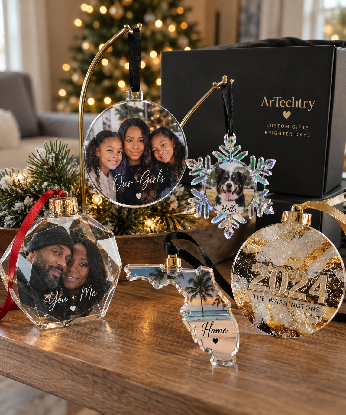 Decorative Christmas ornaments with personal photos and text on a wooden table, with a Christmas tree in the background. The ornaments include a circular photo of a woman and two girls with the caption 'Our Girls', a snowflake shape with a dog and the name 'Bella', a faceted ornament with a couple and the caption 'You + Me', a shape of Texas with a tropical beach scene and the caption 'Home', and a glittery ornament with '2024 THE WASHINGTONS'. A black box with gold lettering and gold ribbon is also on the table.