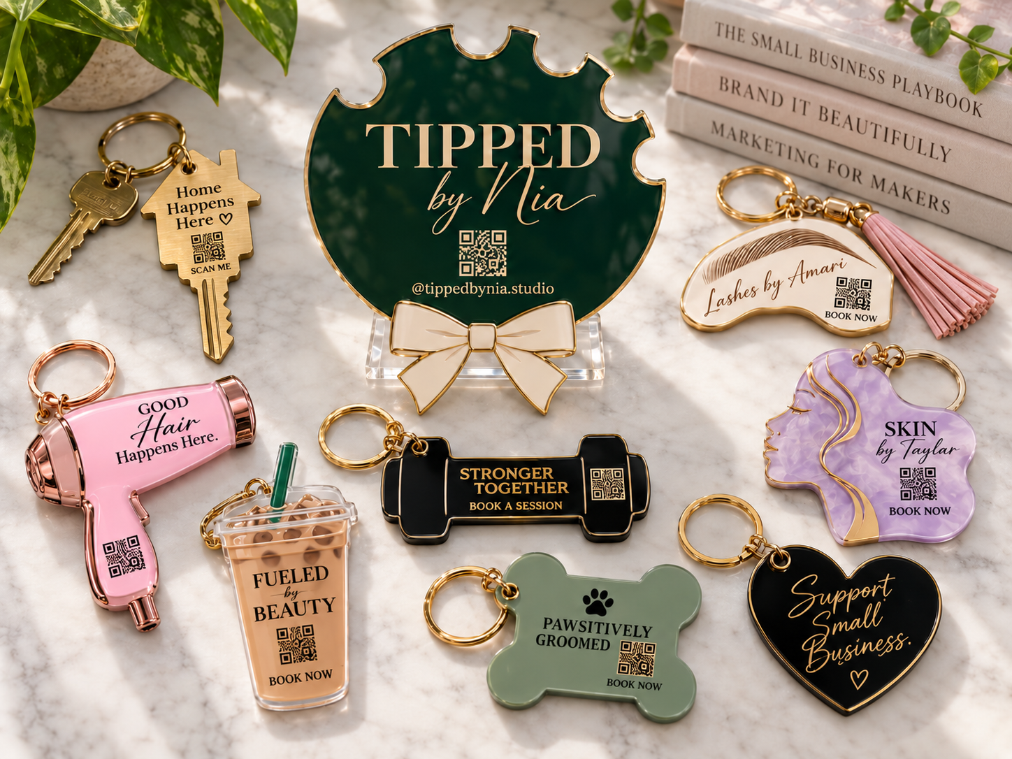 A collection of embroidered and acrylic keychains and ornaments with motivational and business-related messages, including a green sign that says "Tipped by Nia" with a QR code, on a marble surface with books and green leaves in the background.