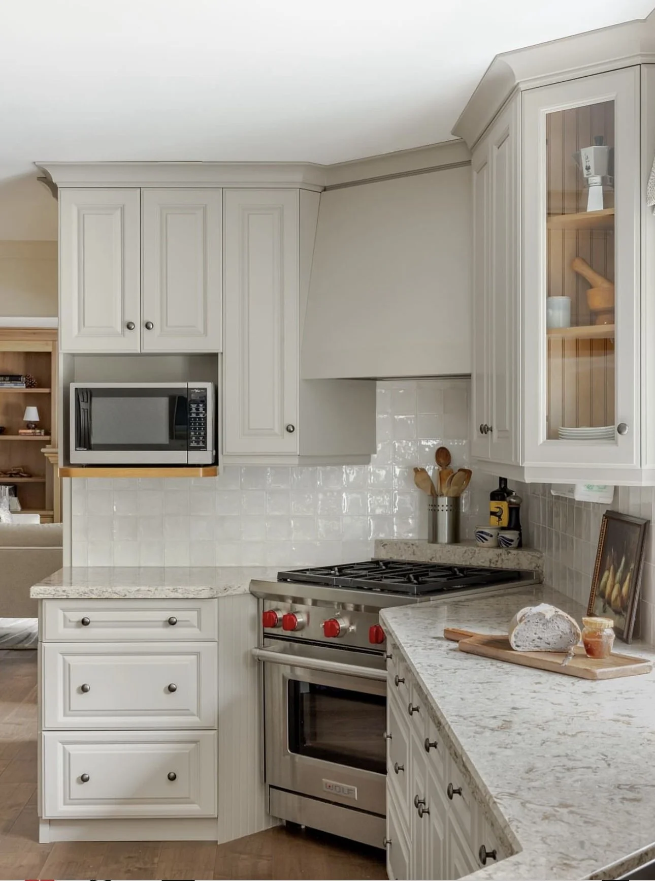 Modern kitchen with white cabinets, a stainless steel oven, microwave, and gas stove. A marble countertop features a cutting board with bread and jars. Decor items are visible on the shelves.