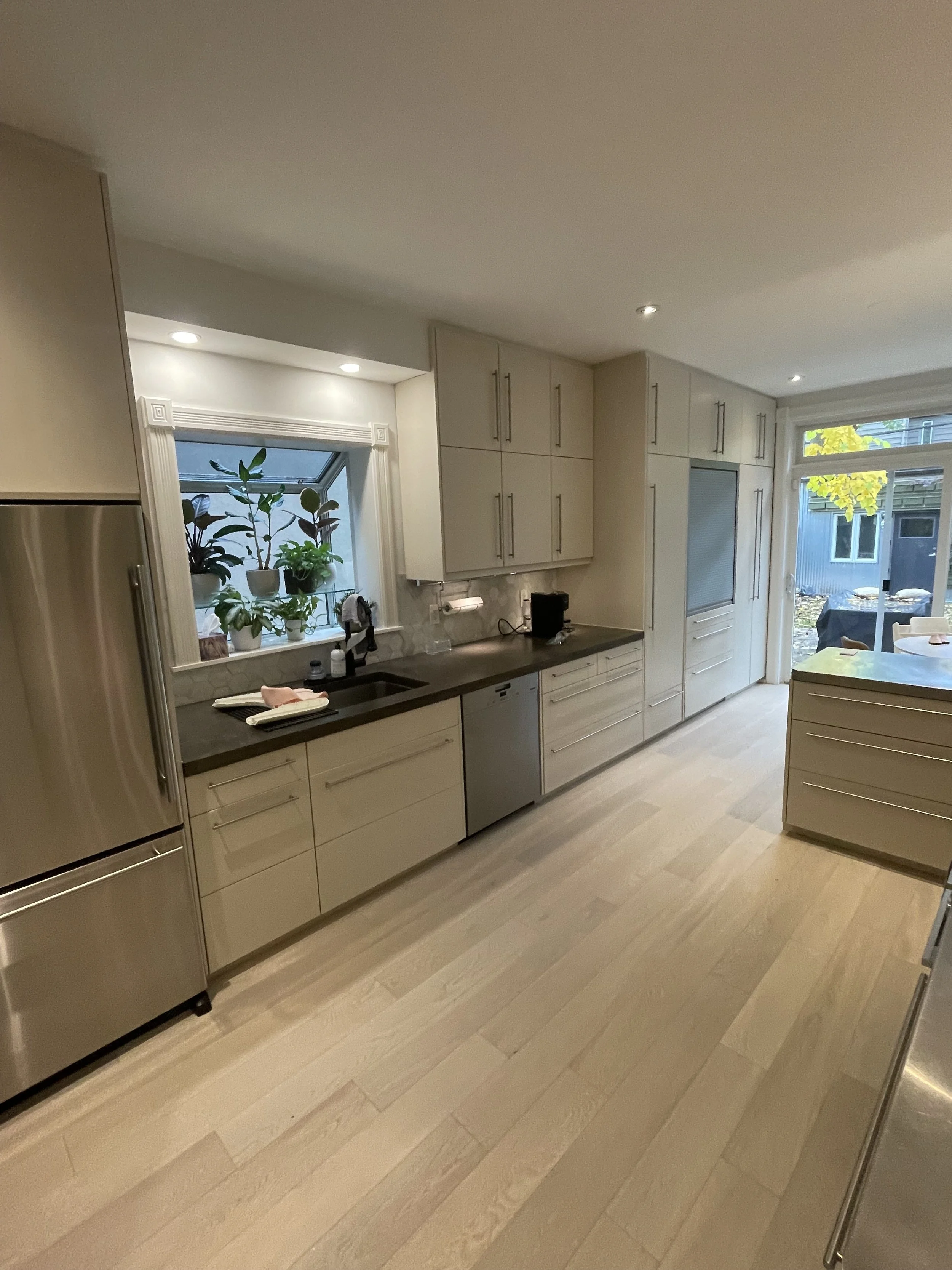 Modern kitchen with light wood flooring, stainless steel appliances, white cabinets, and a countertop with plants by a window.