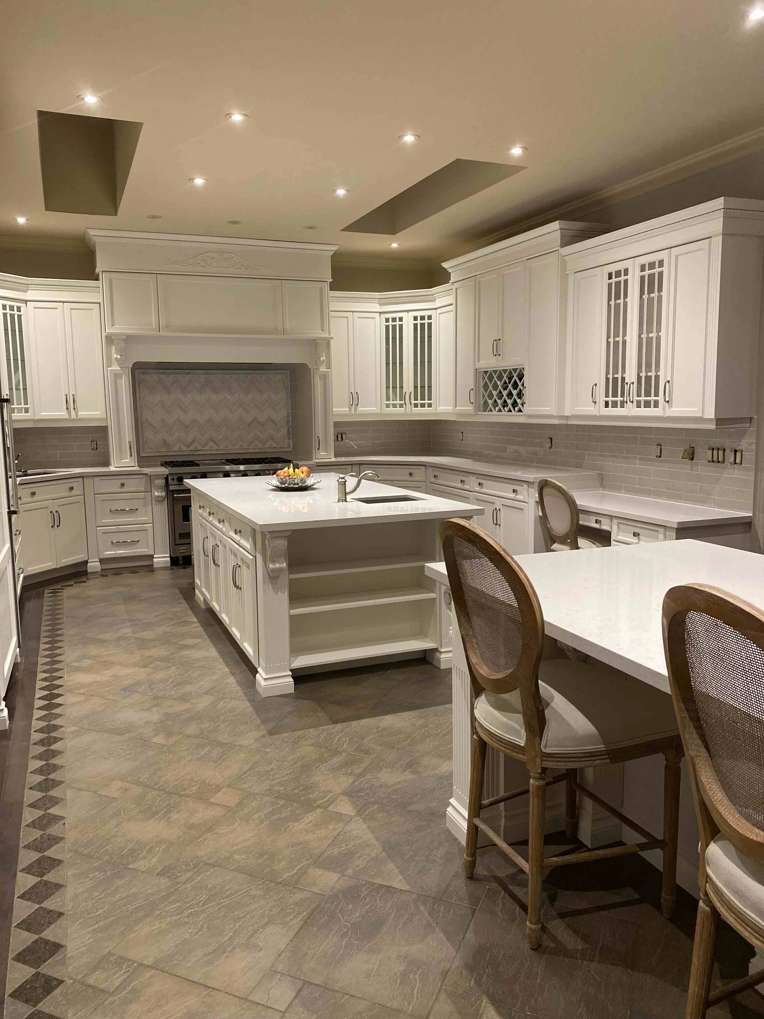Modern kitchen with white cabinets, a large island, high ceilings, recessed lighting, and tiled flooring. There are two cushioned chairs and a bowl of fruit on the countertop.