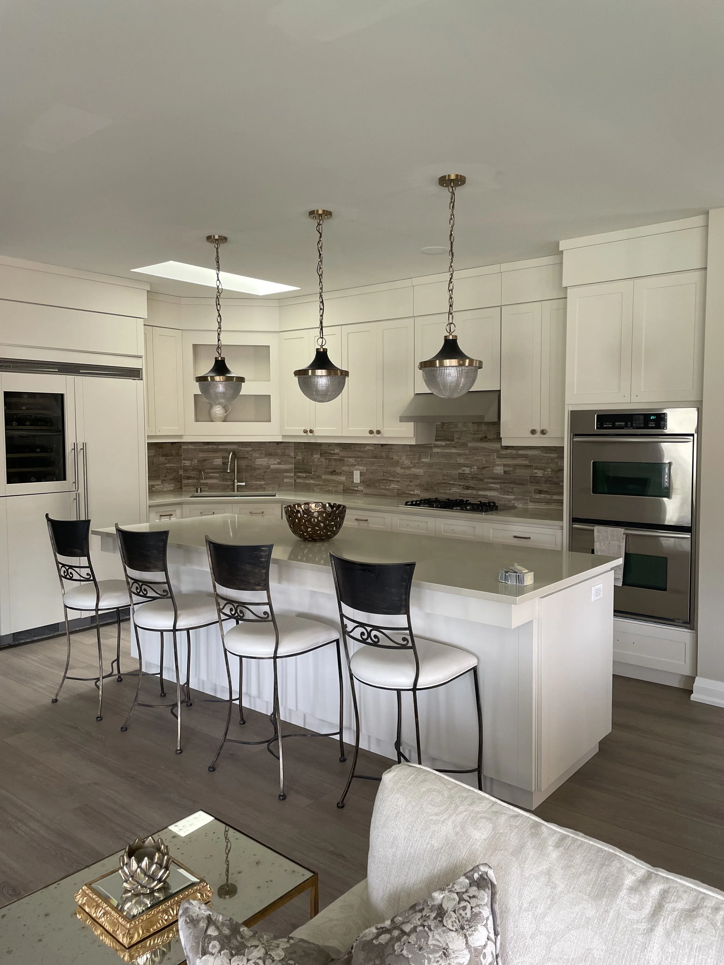 Modern kitchen with white cabinetry, three pendant lights above a large island with bar stools, stainless steel appliances, and a decorative bowl on the counter.