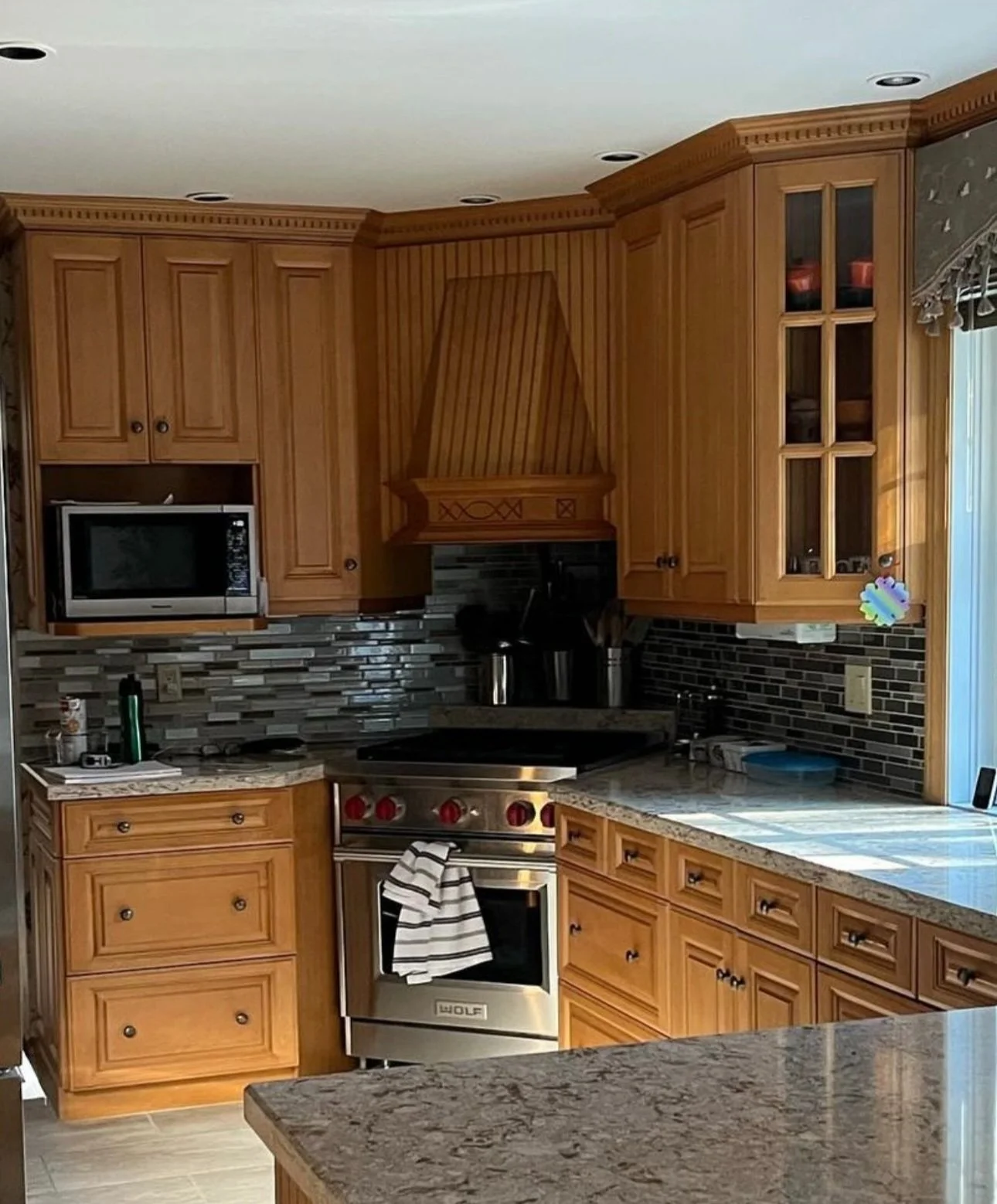 Kitchen with wooden cabinets, granite countertops, a stainless steel stove with a towel hanging on the oven handle, a microwave, a glass-front cabinet, and a tiled backsplash.