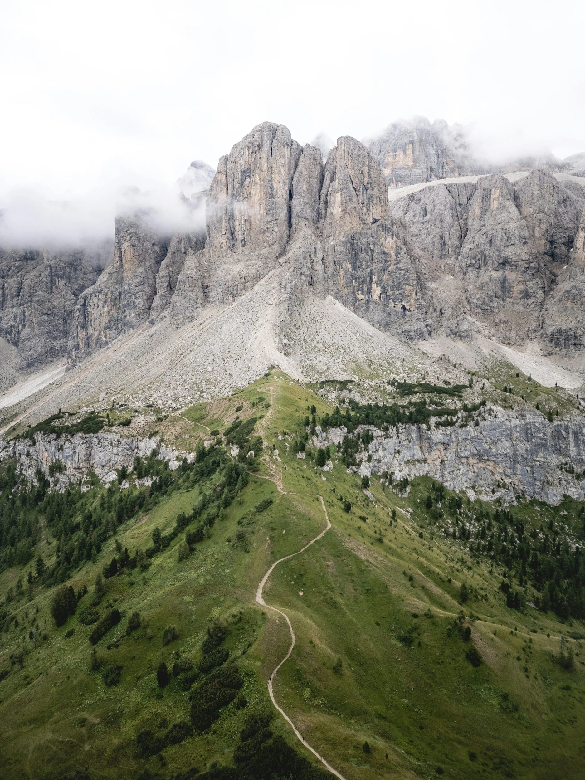 A mountain with steep, rugged rocky peaks partially covered by clouds above a green hillside with a winding dirt trail.