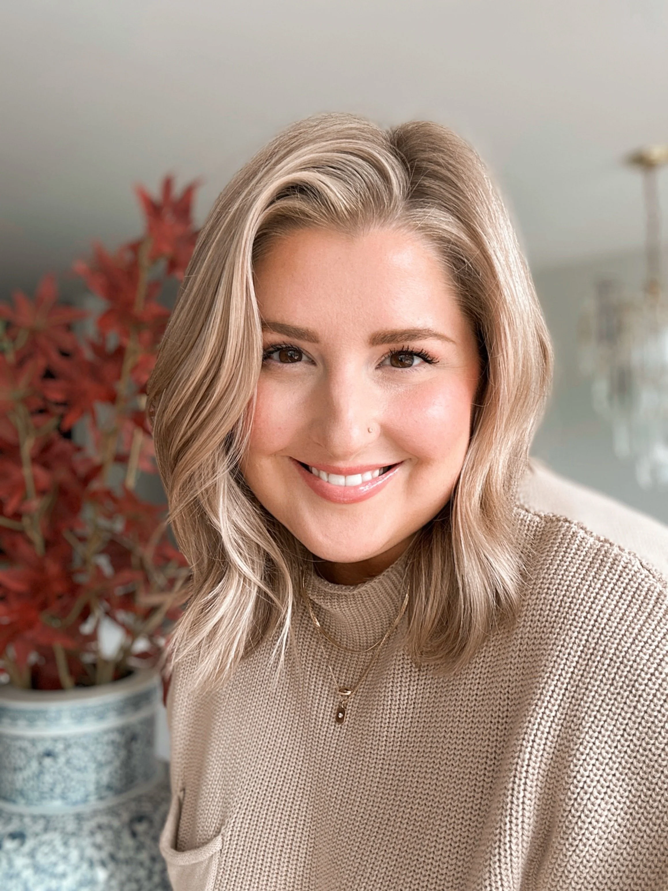 Close-up of a smiling woman with blonde hair and a nose piercing, wearing a beige sweater and layered necklaces, with a potted plant and chandelier in the background.