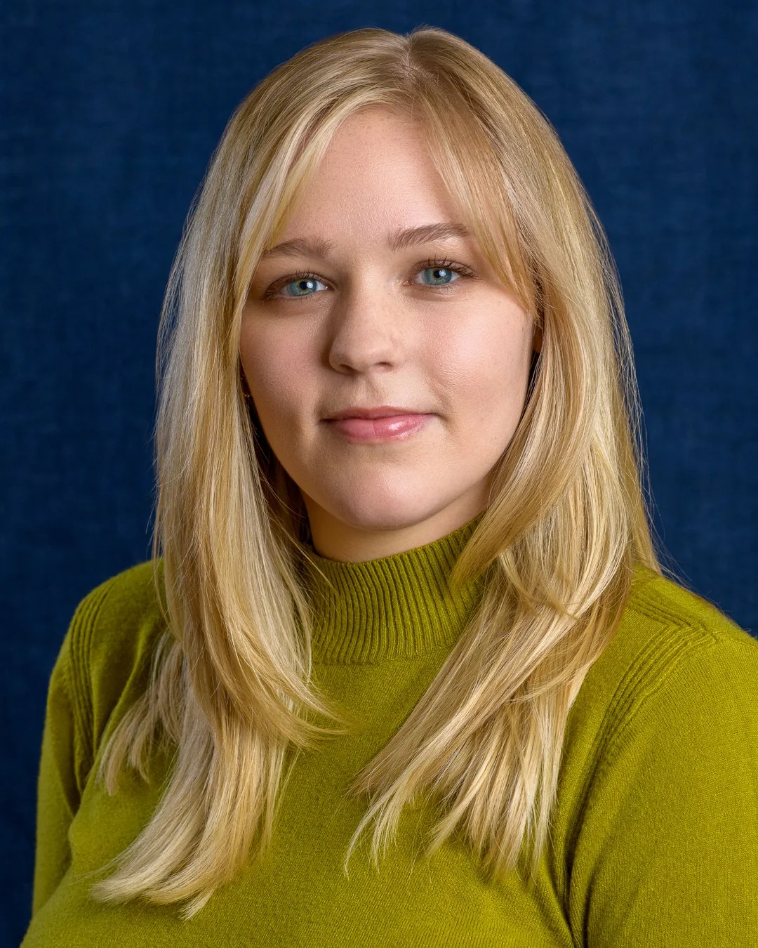 Portrait of a young woman with long blonde hair, blue eyes, wearing a green turtleneck sweater, against a dark blue background.