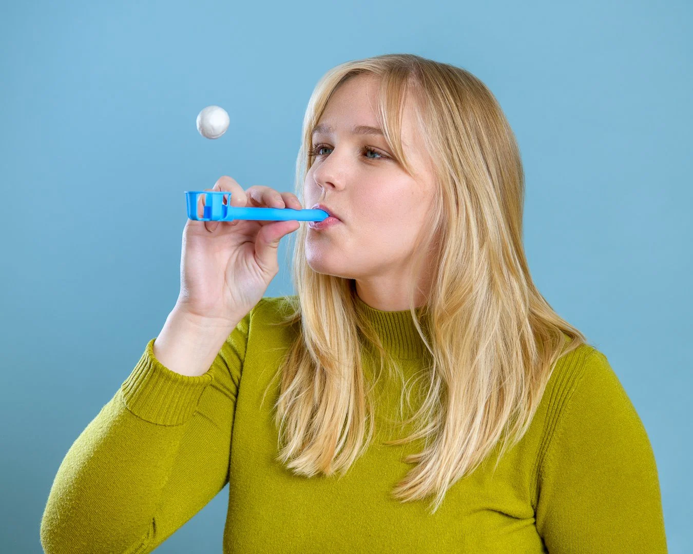 A woman with blonde hair blowing bubbles using a blue bubble blower against a light blue background.